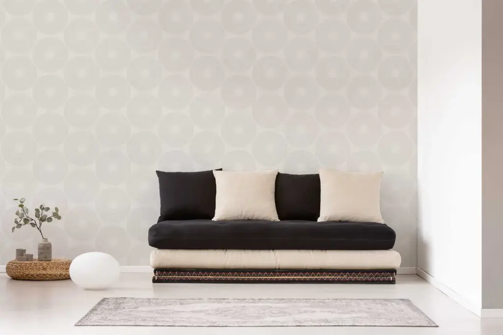 A minimalist living room scene featuring a dark brown sofa with light beige pillows, a grey rug, and a textured white wall.
